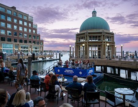 People sit along a waterfront dining area near a domed pavilion by the pier, watching a blue boat perform on the water at dusk.