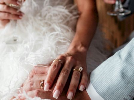 A bride and groom hold hands, wedding rings glow, someone toasting with drinks, feathered dress detail adds soft elegance.