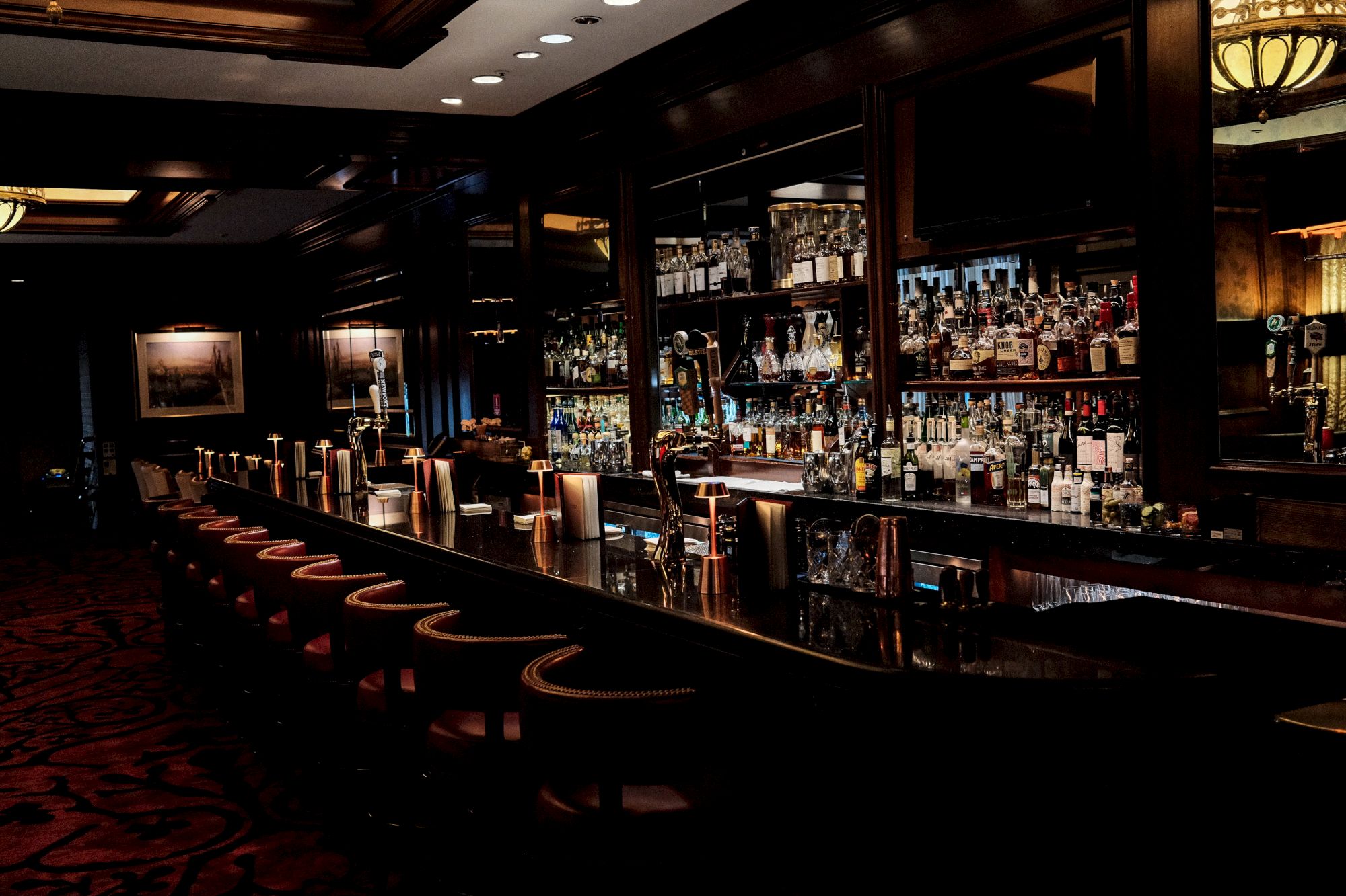 A dimly lit bar with a long counter, lined with stools, shelves of liquor bottles, and classic decor for an elegant atmosphere.