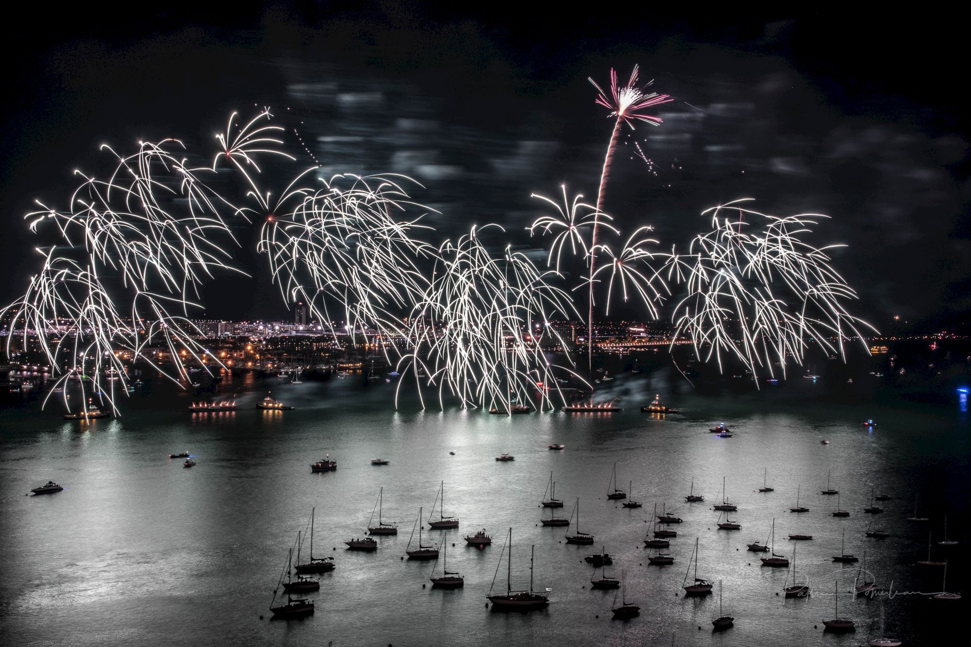Fireworks light up the night sky over a harbor with boats scattered on the shimmering water, creating a festive and vibrant atmosphere.