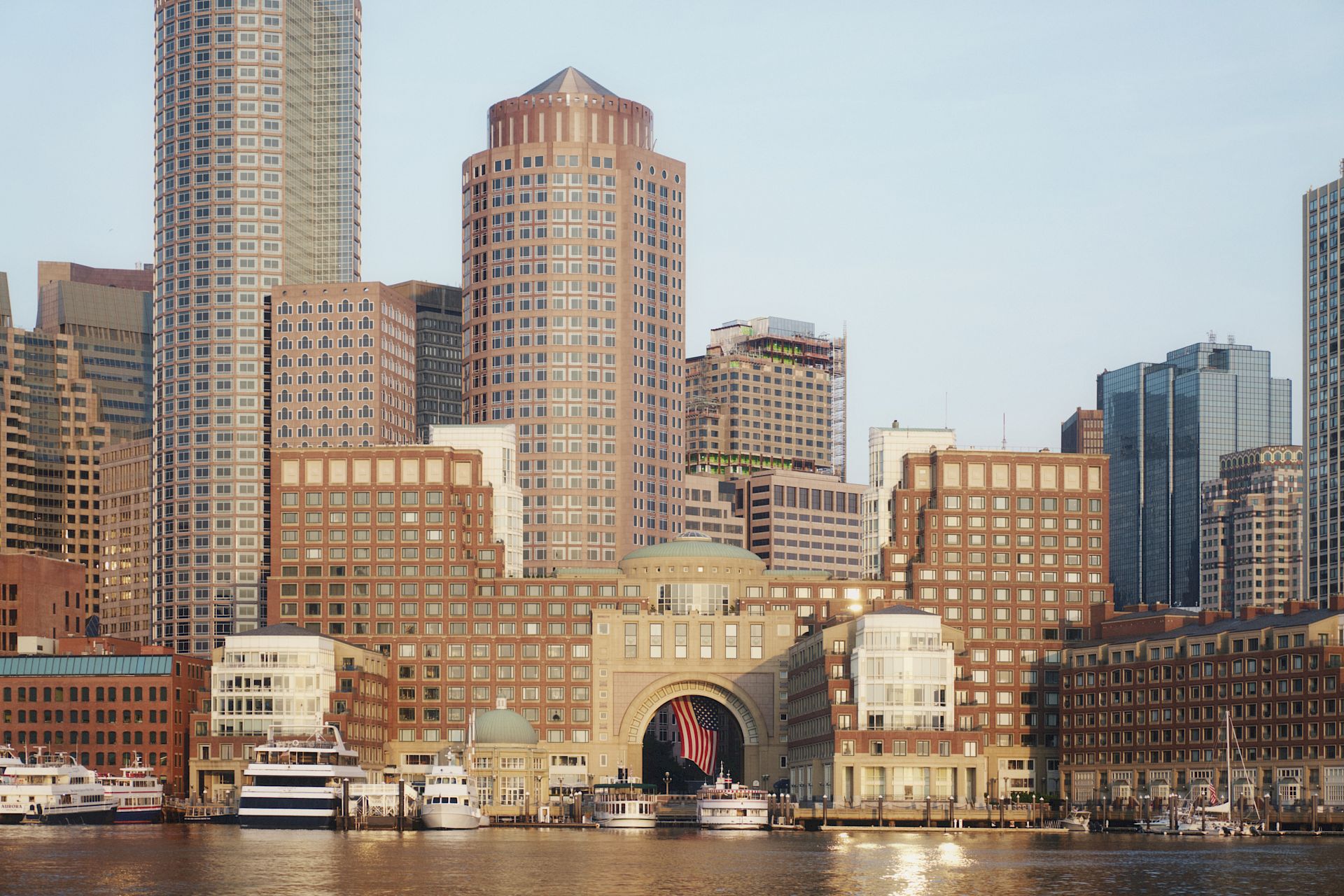 A city skyline of tall brick and glass buildings along a river, with boats docked in the foreground and warm sunset light shining on them.