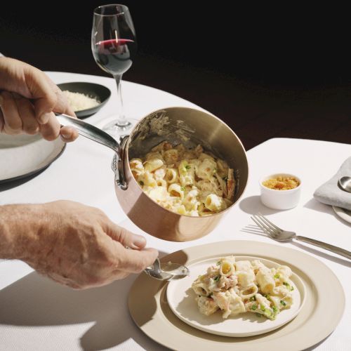 A person serves a creamy pasta dish from a saucepan onto a plated plate, with wine glass and table setting in a fine dining scene.