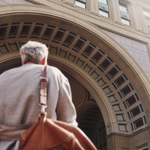 An elderly person with a tan coat and brown bag walks toward a large arched building entrance.