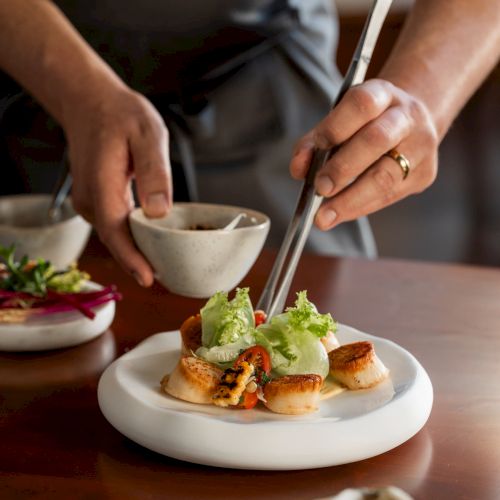 A chef plates a dish of greens and sliced seafood at a dining table, using utensils to serve the food.