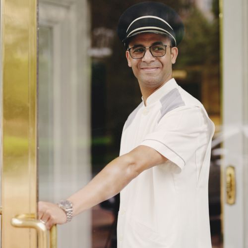 A smiling man wearing a white polo and cap opens glass doors, standing at an entrance.
