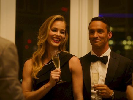 A smiling woman in a black dress raises a champagne flute, chatting with a man in a tux at a lively evening event, clinking glasses.
