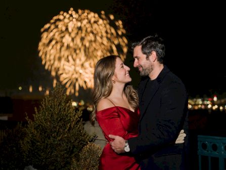 A couple in formal wear shares a tender moment as fireworks sparkle overhead during a night celebration.