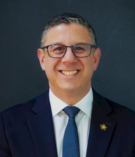 A smiling man in a dark suit, white shirt, blue tie, and black glasses stands against a dark background.
