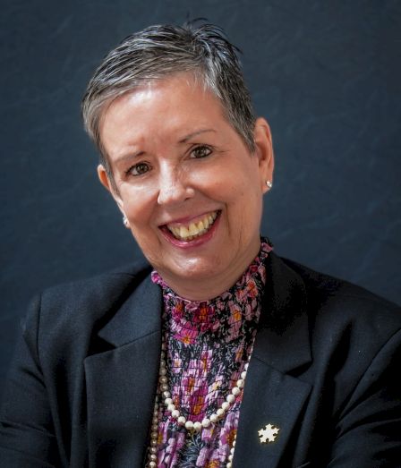 A smiling woman with short gray hair, wearing a dark blazer and layered necklaces, posing against a blue backdrop.