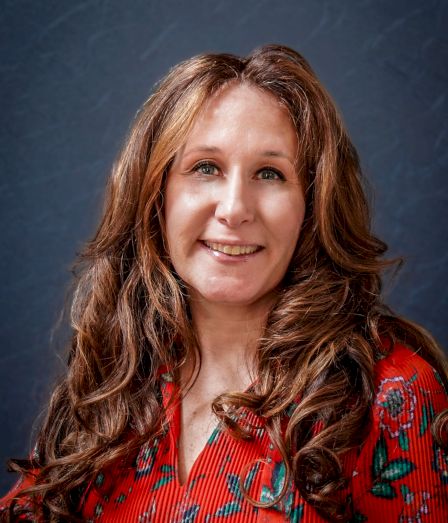 A smiling woman with long wavy brown hair wearing a red floral blouse, posed against a dark blue backdrop.