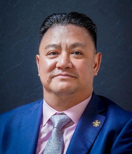 A well-dressed man in a blue suit and pink shirt with a light gray tie, posing confidently against a dark backdrop, looking at the camera.