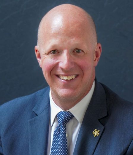A smiling bald man in a blue suit and tie, posing for a portrait against a dark background, wearing a small lapel pin.