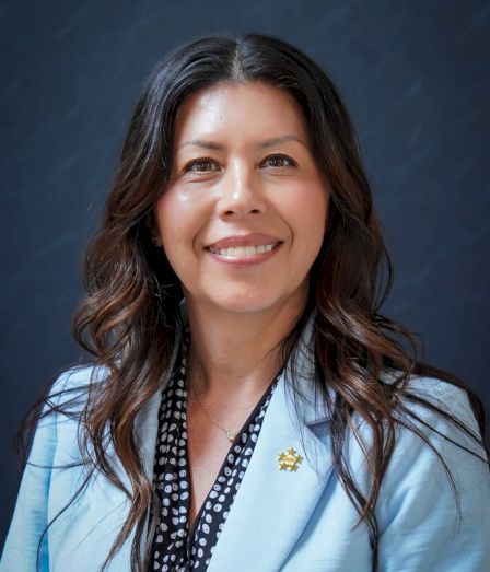 A smiling woman in a light blue blazer and patterned blouse poses for a professional headshot against a blue backdrop.