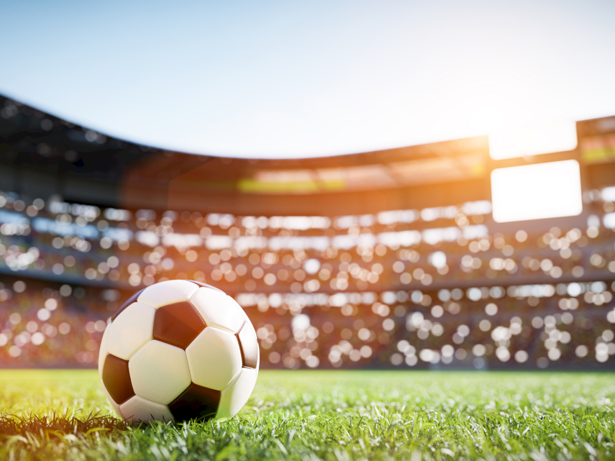A soccer ball on a sunlit stadium field, with a bright crowd and glowing floodlights in the background, under a clear sky.