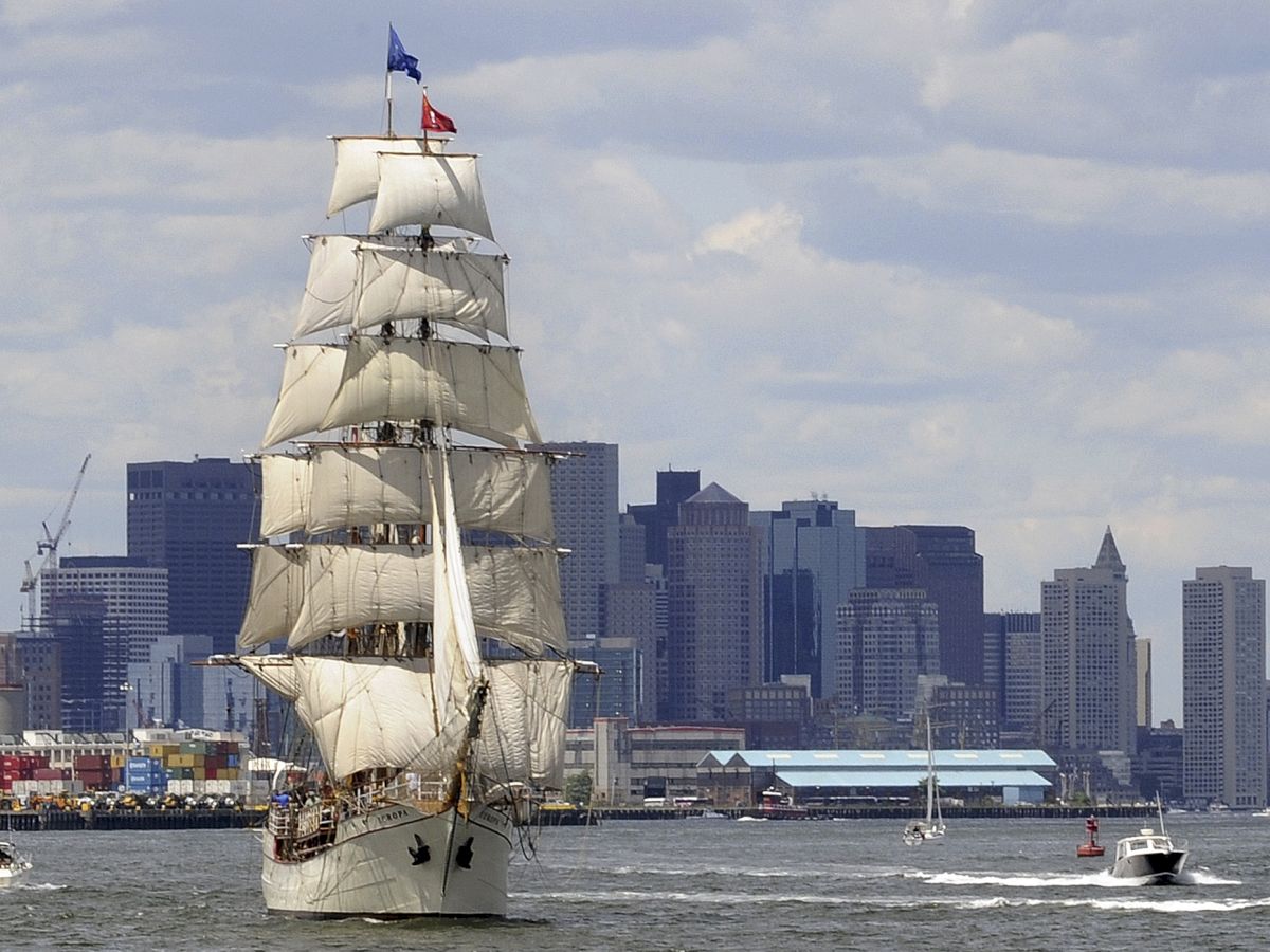 A tall white sailing ship with multiple sails rides near a modern city skyline on the water, with a few smaller boats nearby.