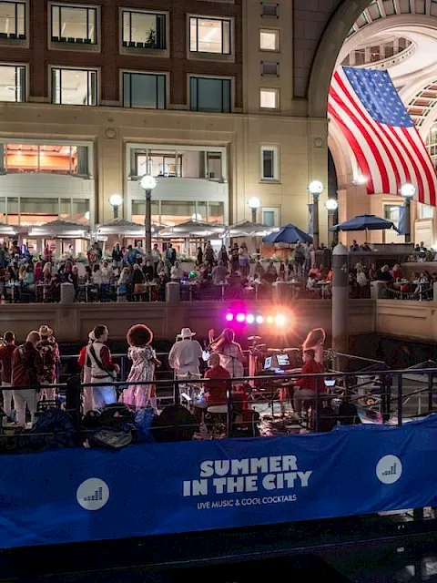 An indoor &ldquo;Summer in the City&rdquo; event with a live band on a stage, a big American flag display, and a cheering crowd.