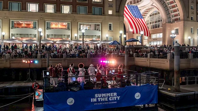 An indoor &ldquo;Summer in the City&rdquo; event with a live band on a stage, a big American flag display, and a cheering crowd.