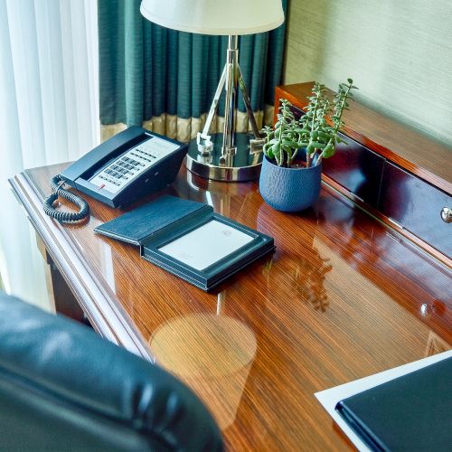 A hotel desk with a corded phone, notebook/journal, lamp, and a small potted plant beside a built-in drawer and window.