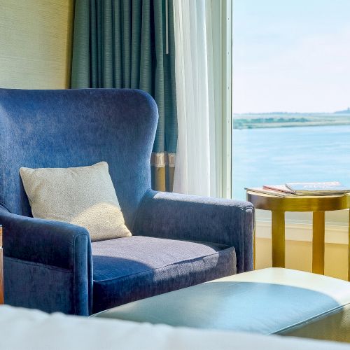 A cozy room with a blue armchair, pillows, a small table with books, and big curtains beside a window overlooking calm water.