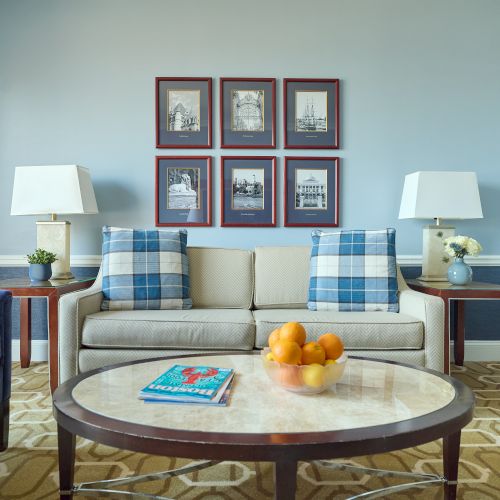 A bright living room with a sofa, coffee table with oranges, two lamps, and six framed pictures on a blue wall.