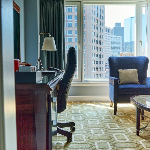 A modern hotel office suite with a blue armchair, desk lamp, rolling chair, table, and city view through large windows.