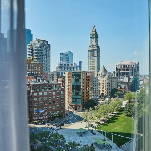 A downtown city skyline with tall buildings and a prominent clock-tower, viewed from indoors under a clear blue sky.