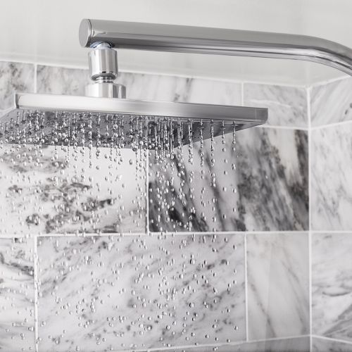A chrome showerhead sprays water over a marble-tiled shower wall, with droplets sparkling in the air.