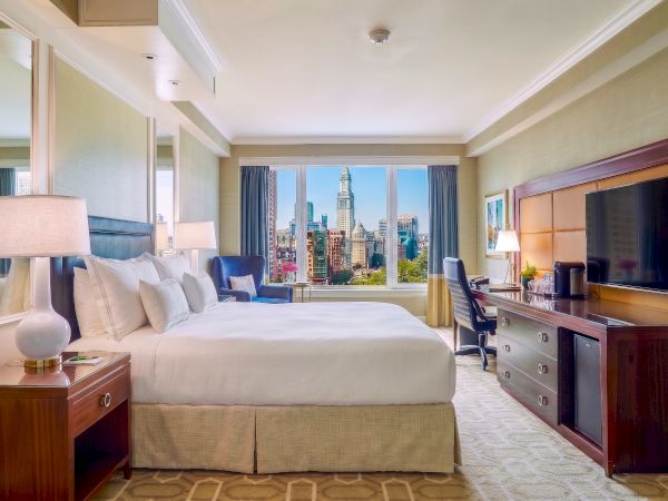 A luxury hotel bedroom with a king bed, lamps, desk, and a city skyline view through large windows.
