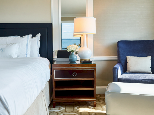 A hotel room with a bed, bedside table, lamp, vase of white flowers, and a blue armchair by a window.