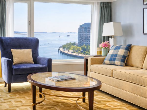 A bright hotel living room with a blue armchair, sofa, round coffee table, and curtains framing a seaside view.