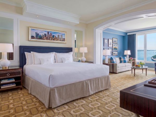 A luxury hotel bedroom with a king bed, lamps, framed art, and a view through large windows over the water.