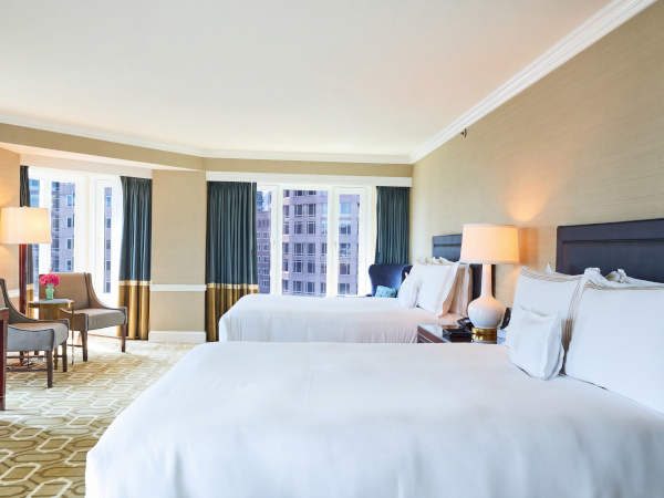 A bright hotel room with two beds, lamps, curtains, and a view of apartment buildings outside, featuring tidy modern d&eacute;cor.