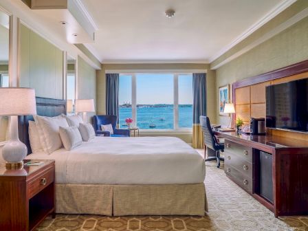 A bright hotel room with a large bed, pillows, lamps, TV, desk, and a window view over the ocean.
