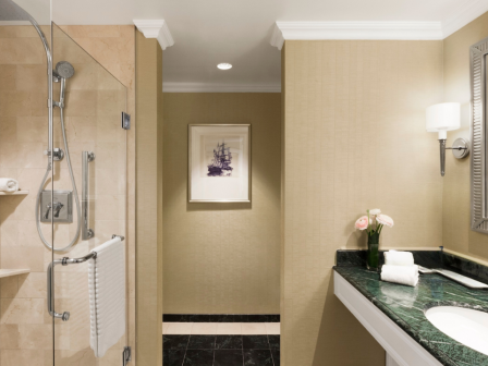 A hotel-style bathroom with a glass shower, green marble sink counter, flowers, towels, and framed wall art in warm lighting.