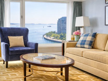 A bright hotel living room with a blue armchair, sofa, round coffee table, and curtains framing a seaside view.