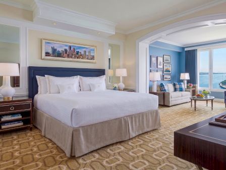 A luxury hotel suite with a neatly made bed, bedside lamps, framed art, and a cozy seating area by large windows.
