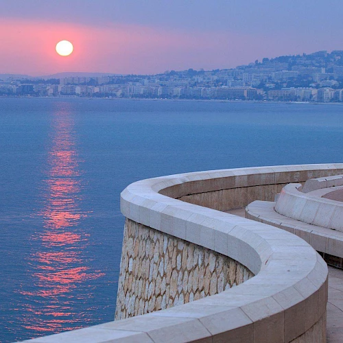 A winding stone pathway curves along a waterfront, with a city skyline and setting sun reflected on the calm sea, under a purple-toned sky.