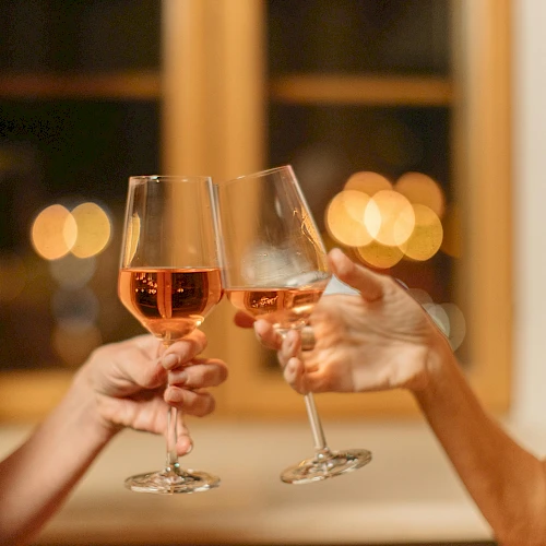 Two people clinking glasses of ros&eacute; wine in a warmly lit setting, with blurred lights in the background creating a festive atmosphere.