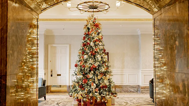 A grand indoor Christmas tree covered in gold and red ornaments with wrapped gifts underneath in an elegant marble lobby.