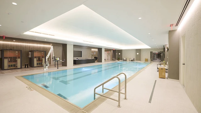 An indoor rectangular swimming pool with clear blue water, modern lighting, long lounge area, and a step ladder at the near end.