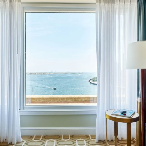 A hotel room with curtained window overlooking a wide river, boats, and a small side table with lamp beside a sofa.
