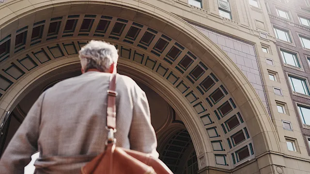 A man walks away beneath a huge arched stone building facade while carrying a guitar case or bag.