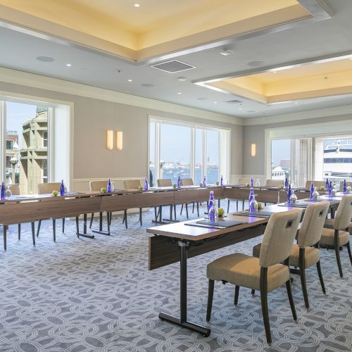 A conference room with tables arranged in a U-shape, chairs, water bottles, and notepads; large windows reveal a waterside view.