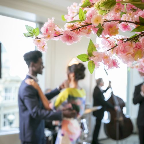 A couple dances in a room with pink blossoms in focus and musicians playing instruments in the background.