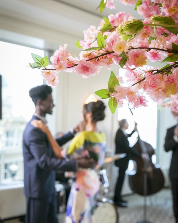 A couple dances in a room with pink blossoms in focus and musicians playing instruments in the background.