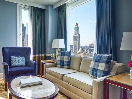 A cozy living room with a sofa, armchair, and a view of a cityscape through large windows, adorned with cushions and a lamp.