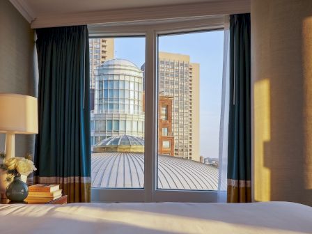 Hotel room window with dark curtains, lamp and books on the bedside table, looking out at city buildings and a glass dome roof.