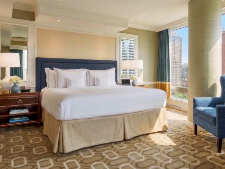 A luxury hotel bedroom with a large bed, lamps, seating, and tall windows looking out on a city skyline.