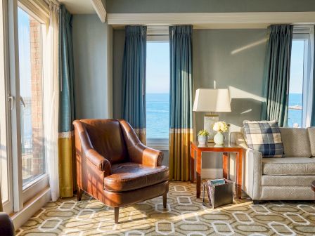 A bright hotel room with ocean-view windows, teal curtains, a leather chair, sofa, side table with lamp, and flowers.