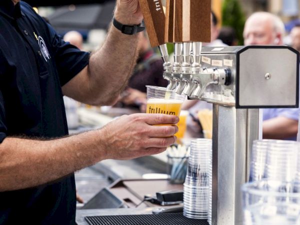 A man pours a drink sample into a small cup from a stainless dispensing machine at a trade-show booth.